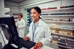 Cheerful young female pharmacist wearing labcoat working on computer in drugstore