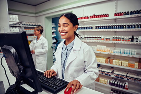 Cheerful young female pharmacist wearing labcoat working on computer in drugstore